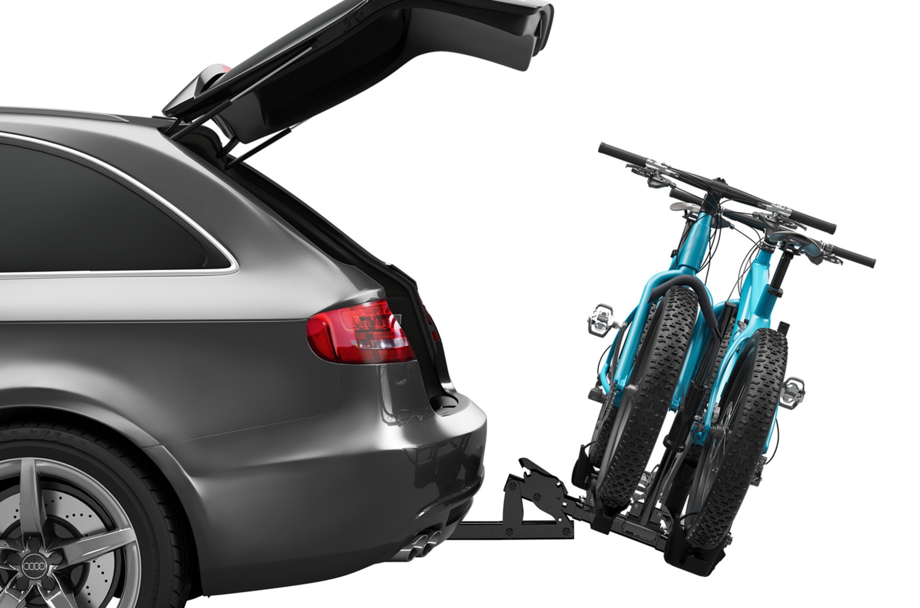 Two blue fat-tire bikes secured on a hitch-mounted bike rack attached to the rear of a gray Audi wagon with the hatch open