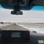 View from inside a van driving on a straight highway through a snow-dusted desert landscape with a GPS navigation screen on the dashboard