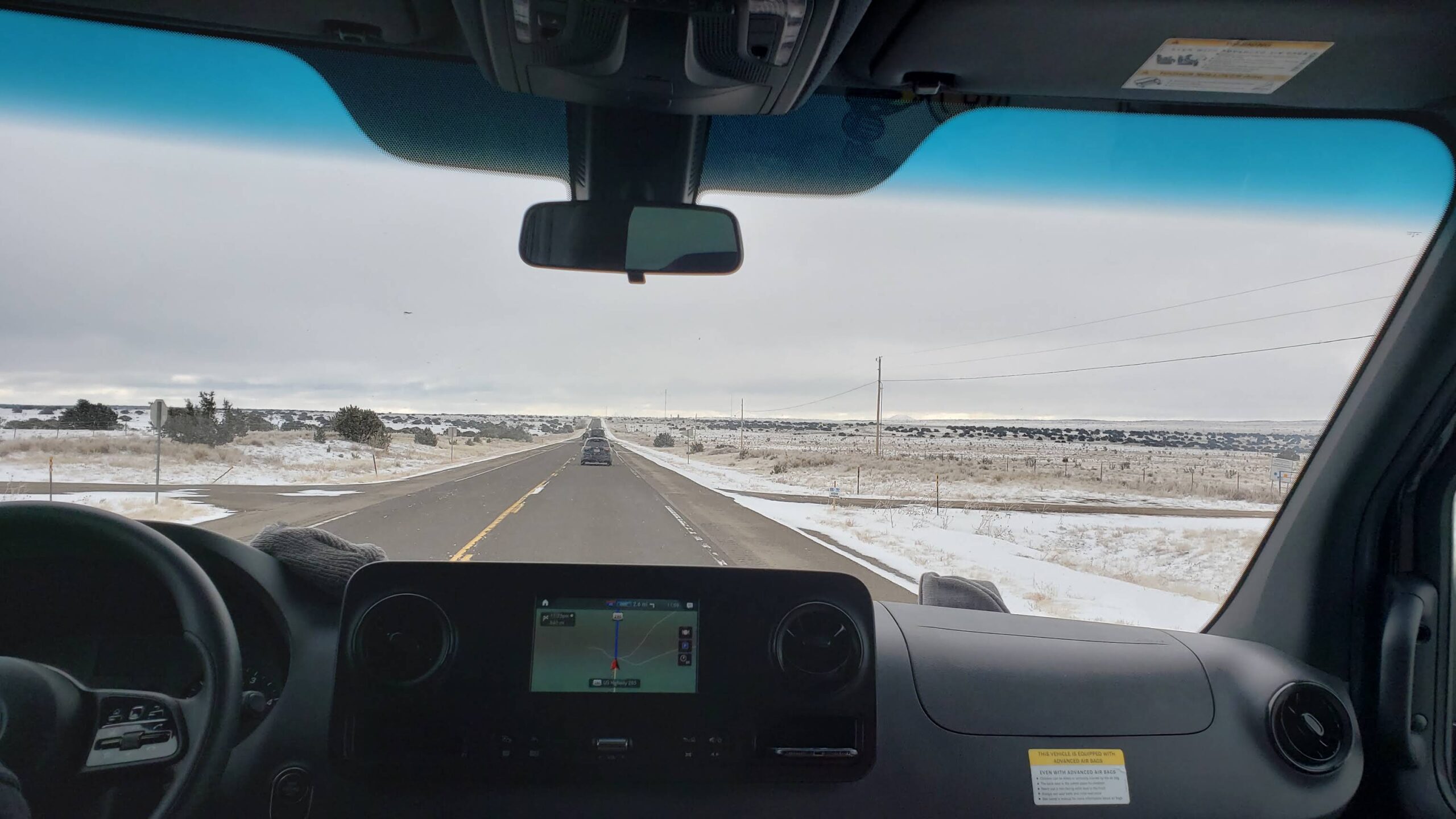 View from inside a van driving on a straight highway through a snow-dusted desert landscape with a GPS navigation screen on the dashboard