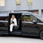 Woman in a white suit sitting in the open sliding door of a black luxury passenger van outside a modern building