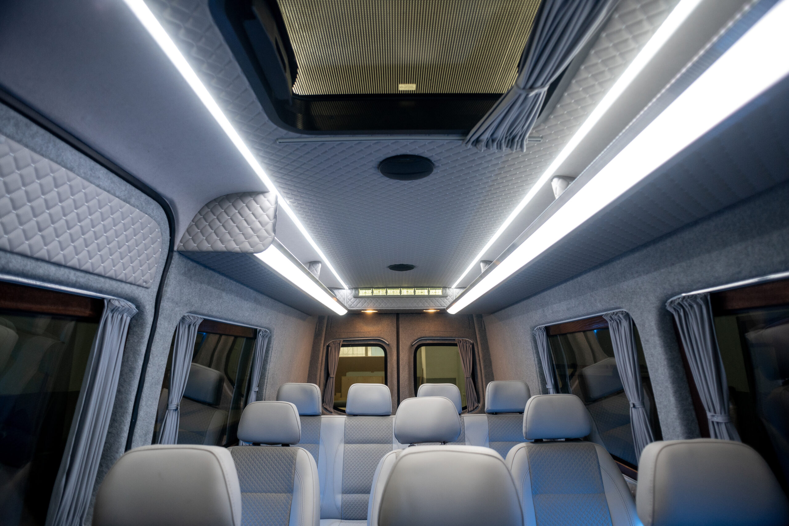 Interior of a luxury passenger van with rows of gray leather seats, LED strip lighting, quilted wall panels, and curtained windows